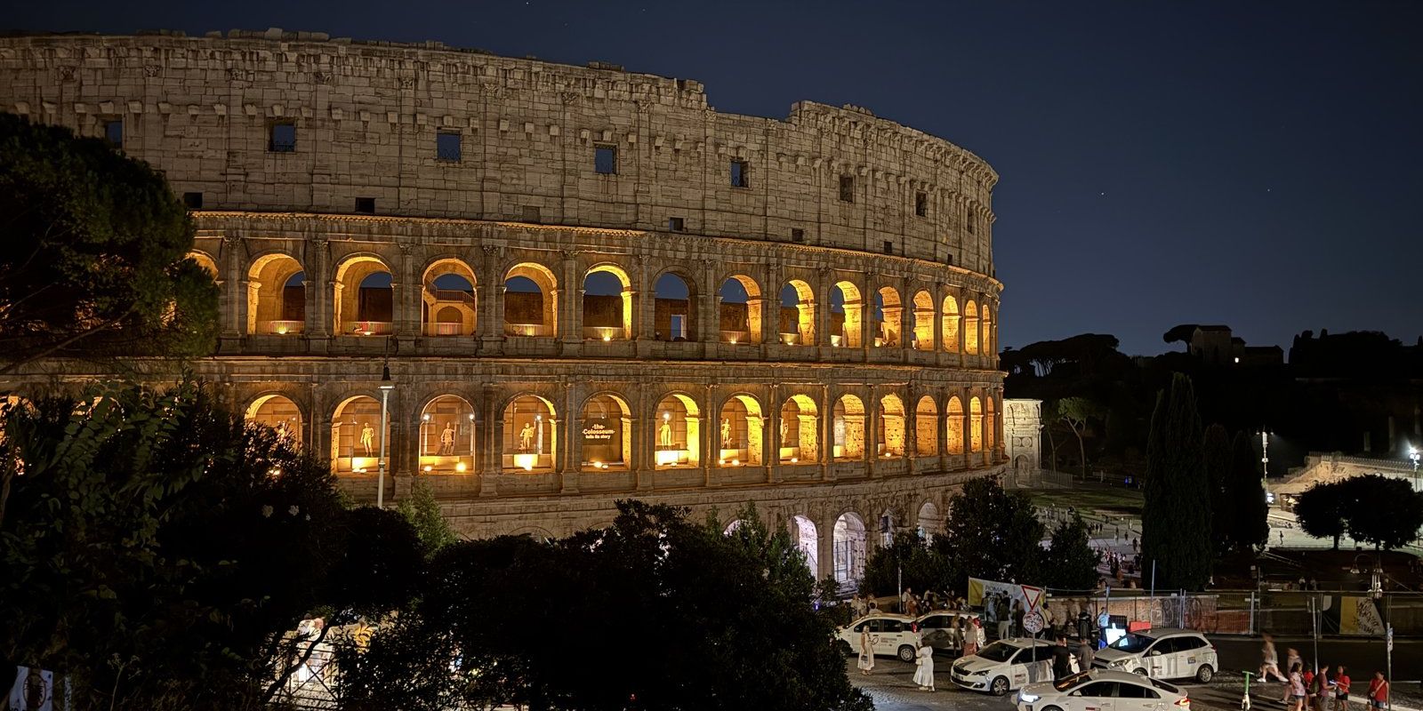 The Colosseum at night, its arches lit from inside, seen from the Oppian Hill side