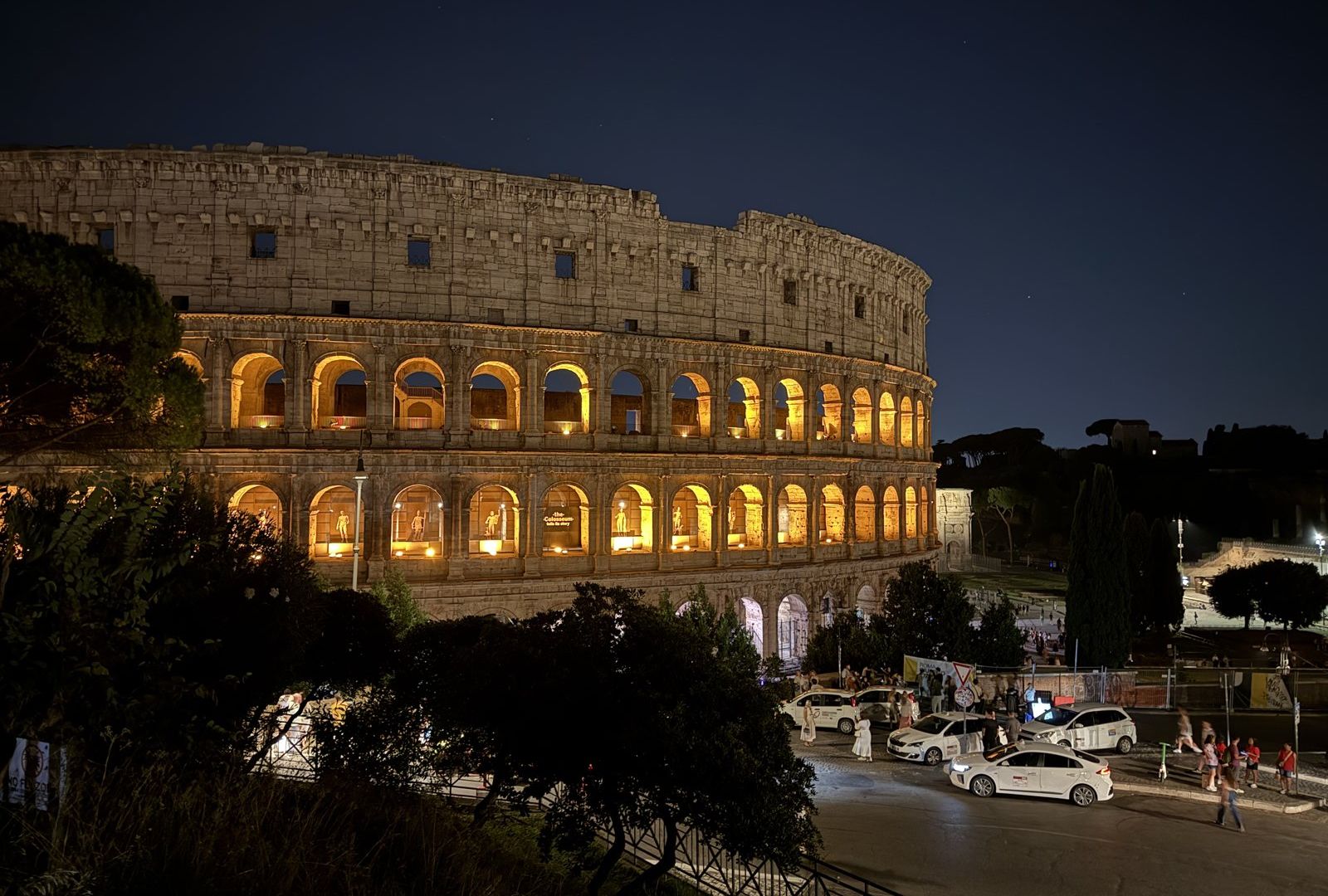 The Colosseum at night, its arches lit from inside, seen from the Oppian Hill side
