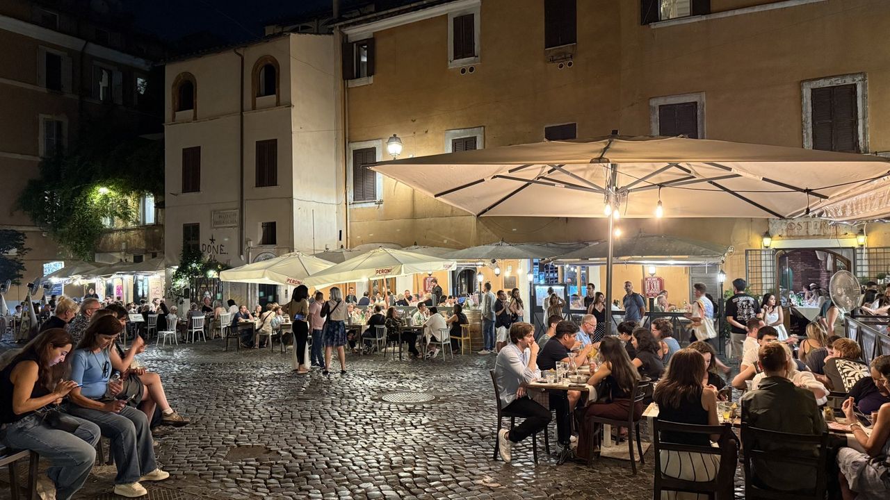 A small Trastevere piazza at night, restaurant tables under umbrellas on the cobbles, people everywhere