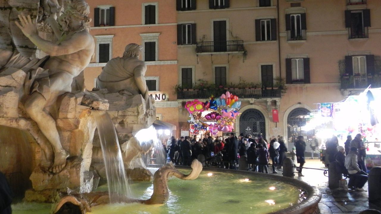 Piazza Navona at night, with the lit Fountain of the Four Rivers, crowds by market stalls, and a vendor's balloons.