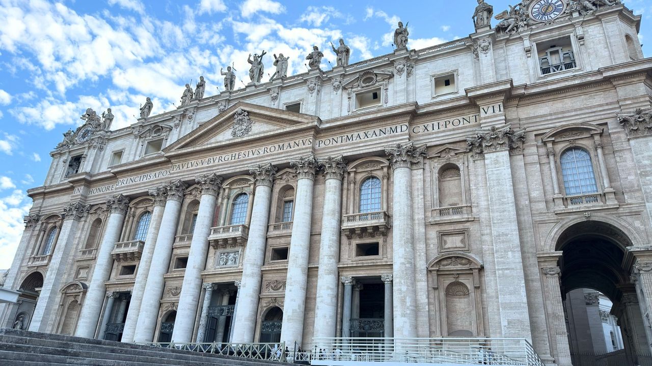 The facade of St Peter's Basilica from below, with its Latin inscription, roofline statues, and barriers on the steps.