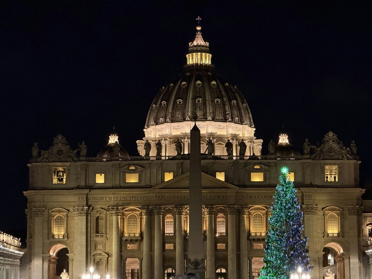 St Peter's Basilica at night behind the obelisk, with a tall Christmas tree in blue and white lights topped by a green star.