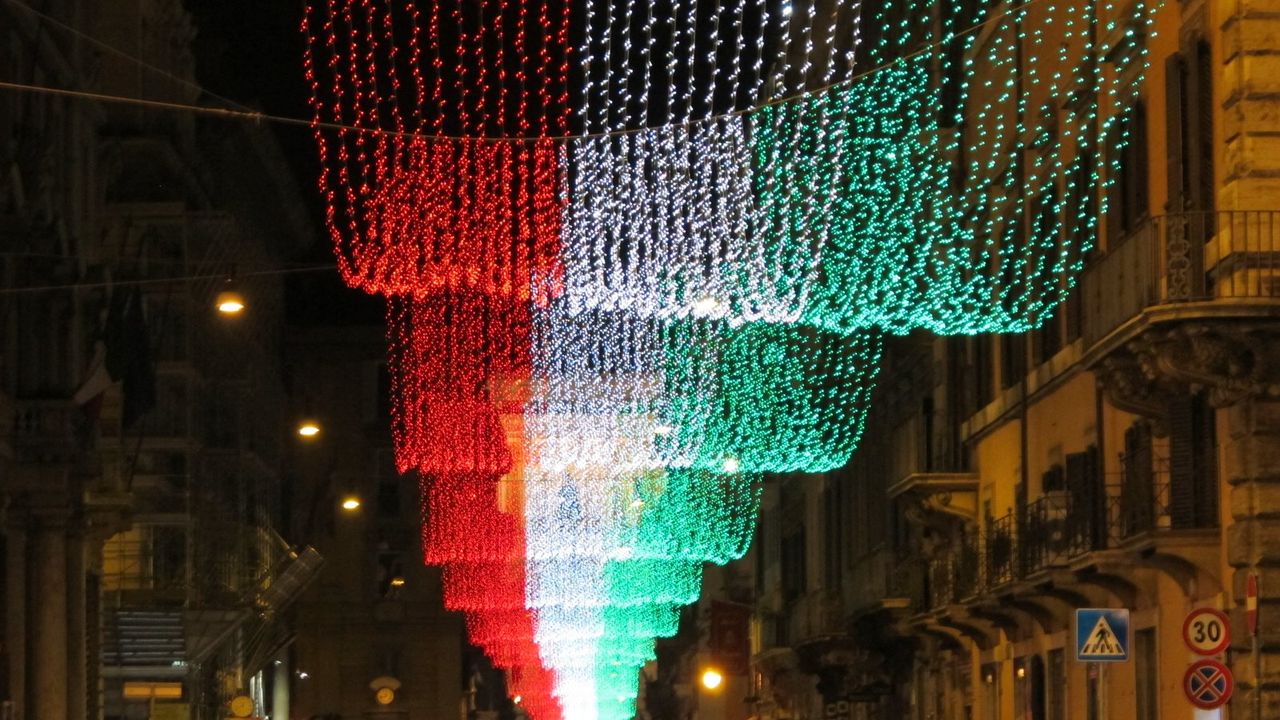 Via del Corso at night with strings of red, white, and green lights cascading down the street toward the far end, cars below.