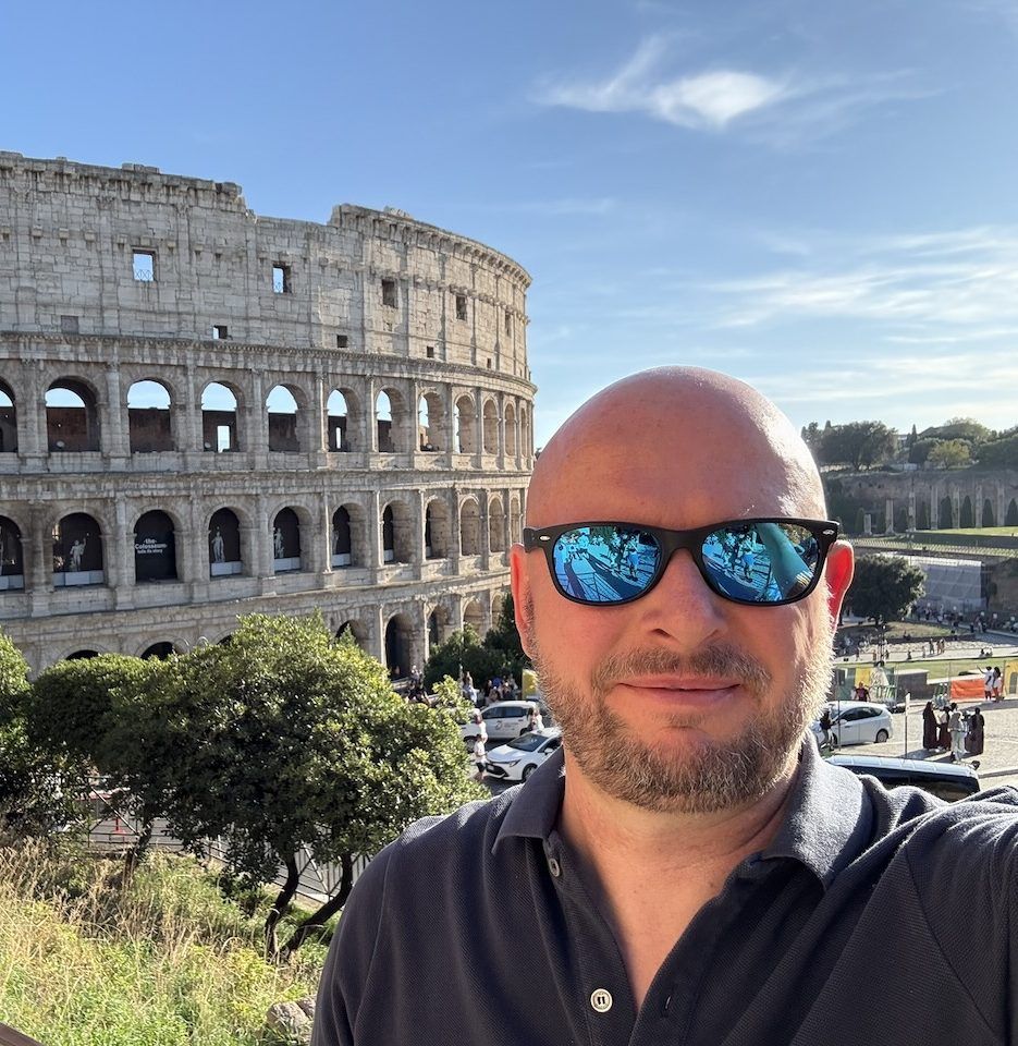 Max in front of the Colosseum on a sunny afternoon, sunglasses on, the arches rising behind him