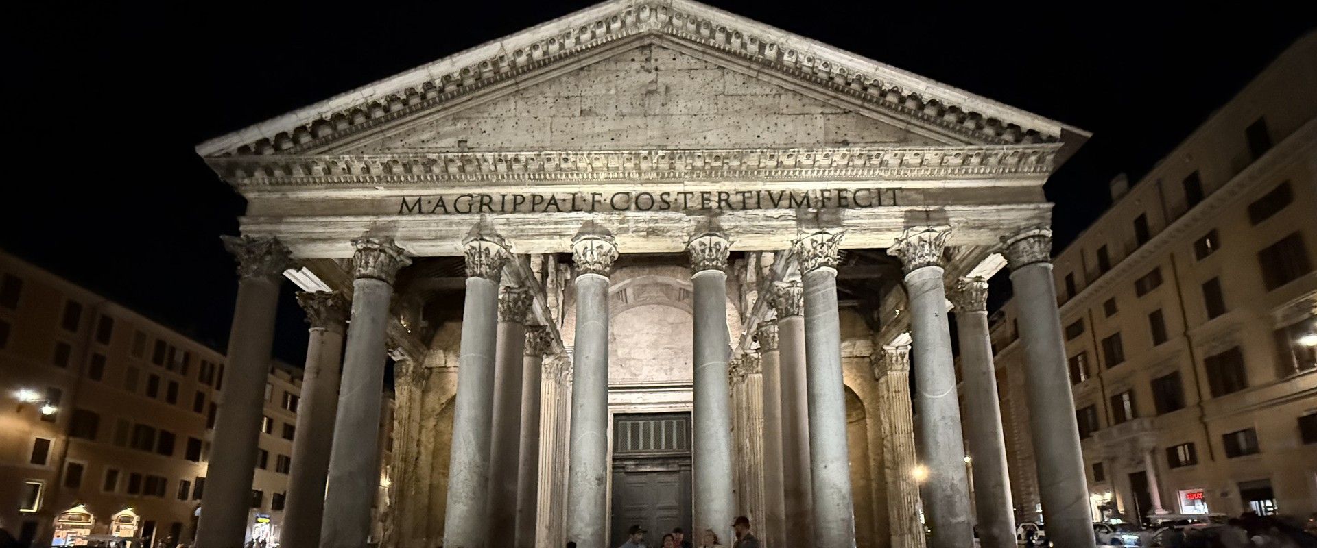 The Pantheon's facade at night, the M. Agrippa inscription lit above the portico columns, people crossing the piazza below