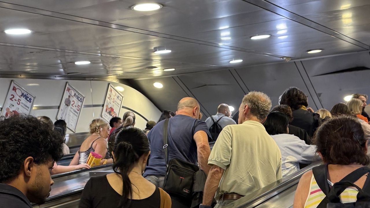 A packed metro escalator in Rome seen from behind, riders pressed shoulder to shoulder with bags and backpacks