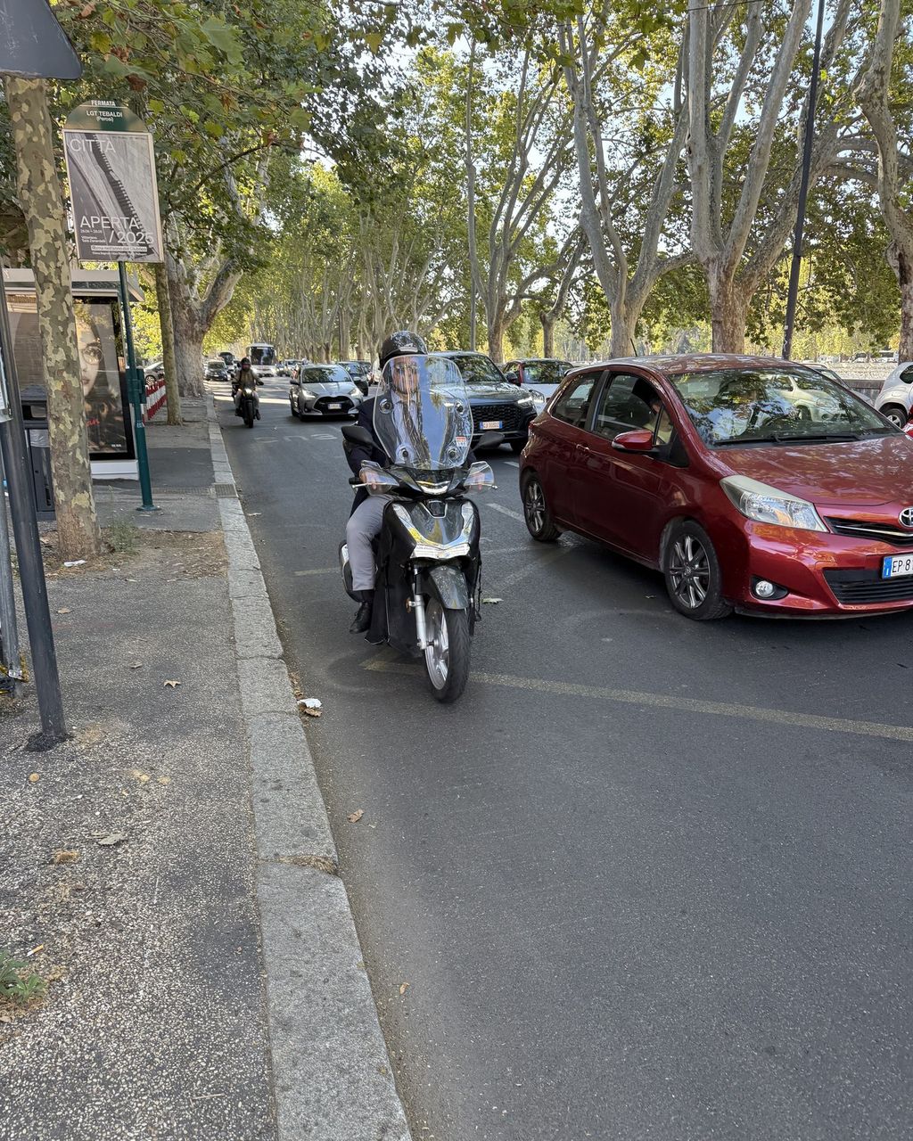 A scooter riding tight against the kerb on a tree-lined Rome street, cars queuing alongside