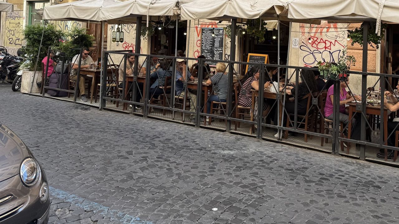 A tourist-zone restaurant terrace in Rome, diners at tables under white umbrellas behind a railing