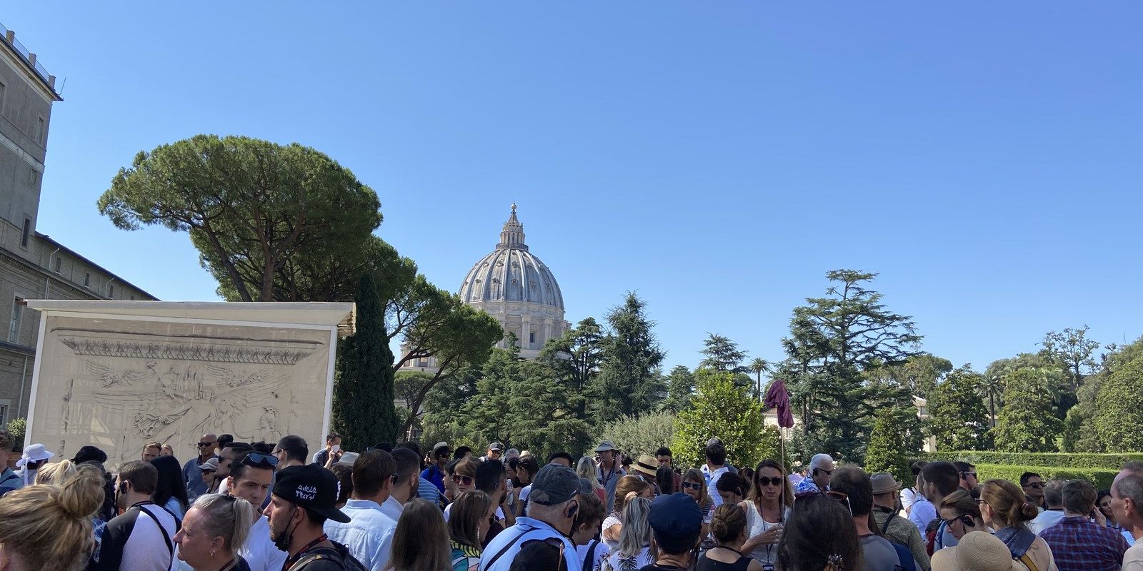 A dense crowd of visitors in the Cortile della Pigna inside the Vatican Museums, St. Peter's dome rising behind the trees