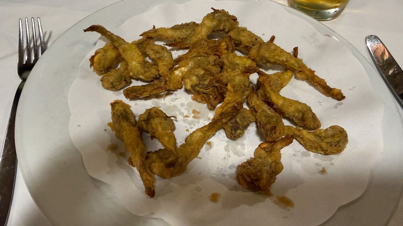 Fried artichoke pieces in golden batter on a paper doily on a glass plate, with a fork and knife on a white tablecloth.