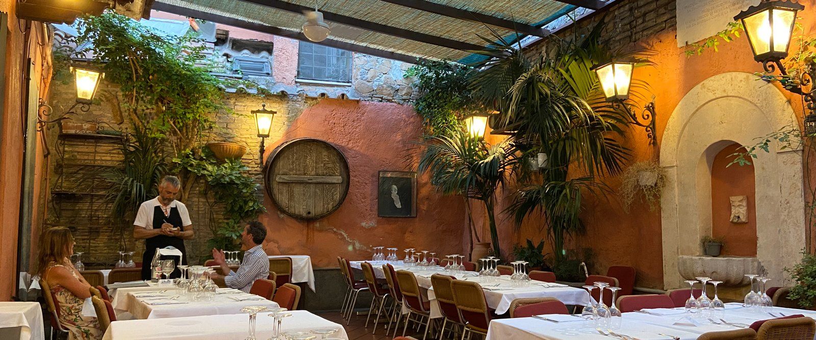 Checco er Carettiere's covered courtyard at evening: linen tables, orange-pink walls, lit lanterns, and a reed-mat roof.