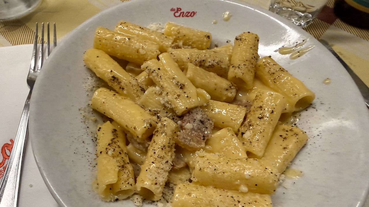 Rigatoni carbonara on a plate printed with the da Enzo logo, topped with pepper, pecorino, and guanciale.