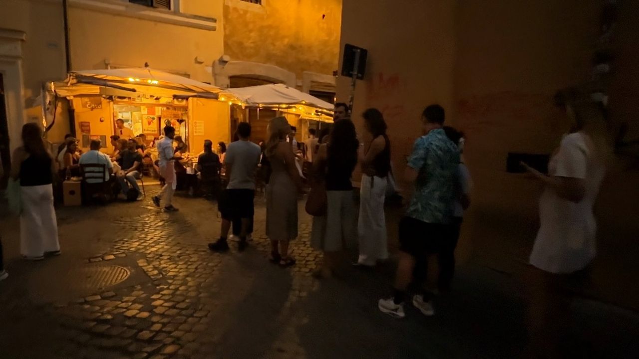 A line of people waits at dusk outside Da Enzo al 29, toward a lit terrace with white awnings and seated diners.