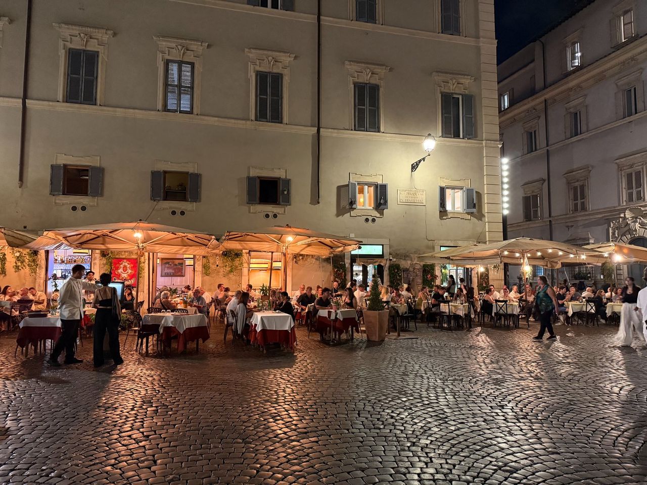 Piazza di Santa Maria in Trastevere at night: lit terraces with red-and-white tablecloths, large umbrellas, and diners.
