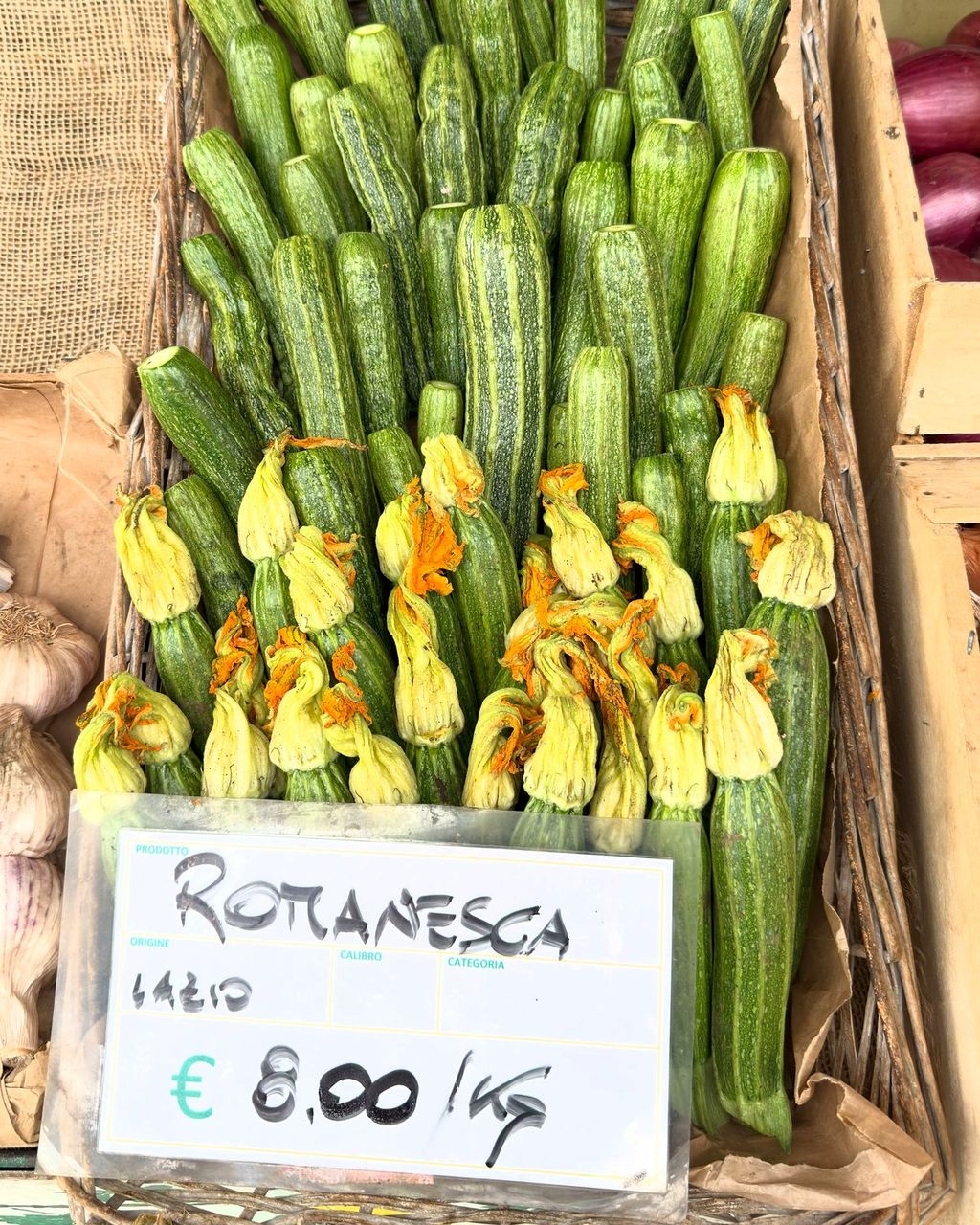 A basket of Romanesca zucchini with orange flowers beside a card reading Romanesca, Lazio, €8.00/kg, and garlic.
