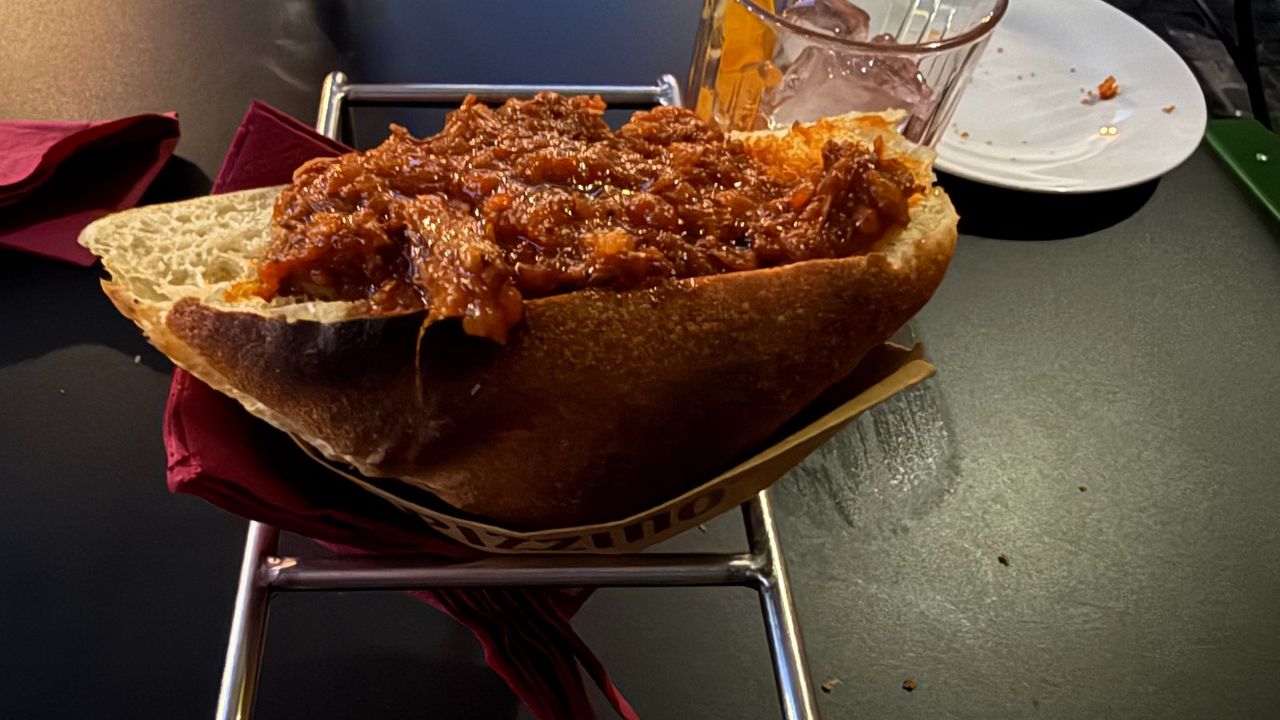 A triangular trapizzino filled with red slow-cooked meat sauce, held in a paper sleeve on a small metal stand.