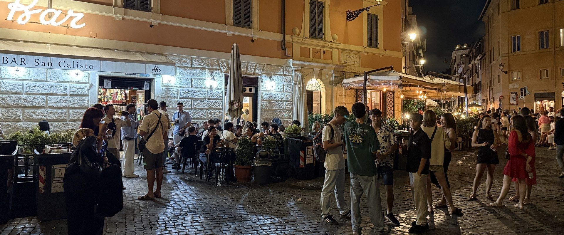 The crowd outside Bar San Calisto at night, drinks on the cobbles of the piazza, the bar's neon sign above