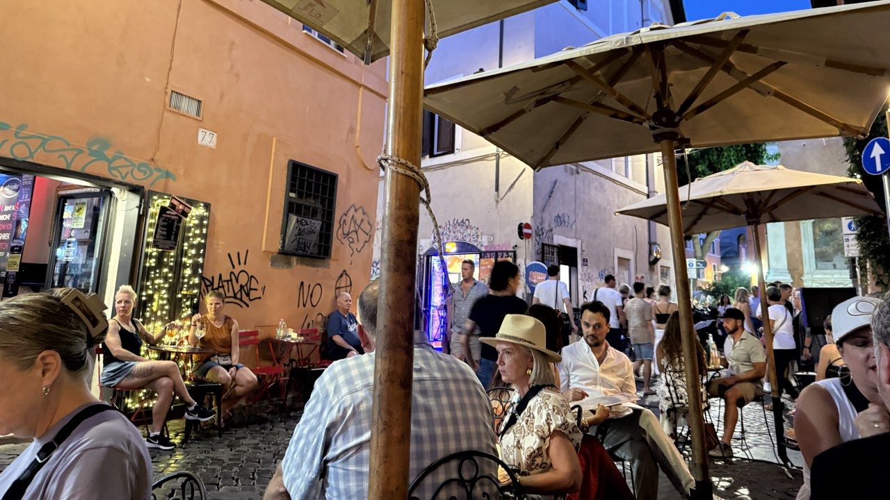 Terrace umbrellas and full tables filling a Trastevere lane at blue hour, string lights on the orange wall